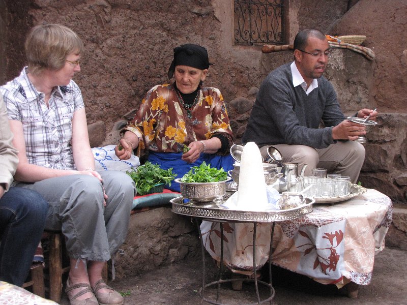 180312_oma fatima muntthee maken.jpg - Na een mooie rondrit komen we bij de tweede Berberse familie uit. We gaan bij de oma thee drinken. Op de foto zit oma Fatima naast Frouwkje. Onze gids Mustapha zit rechts en legt de Berberse traditie van muntthee drinken uit. Zoals je ziet maakt Fatima de thee van verse muntblaadjes.